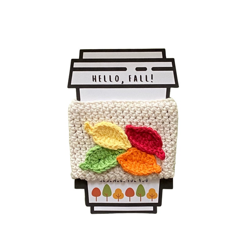 May include: White crocheted coffee cup cozy with a design of five colorful leaves. The cozy has the text "HELLO, FALL!" on the top.
