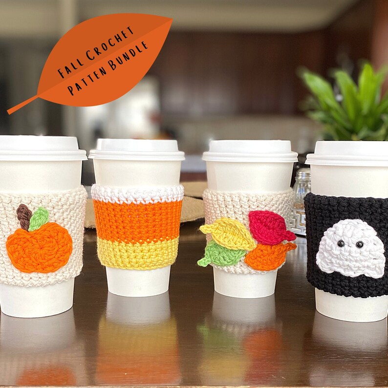 May include: Four crocheted coffee cup cozies in different fall and Halloween themes. The cozies are white, orange, yellow, green, red, and black. The cozies feature a pumpkin, candy corn, leaves, and a ghost.