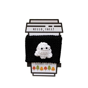 May include: Black and white crocheted coffee cup cozy with a ghost design. The cozy has the text "Hello, Fall!" and "Handmade for you" printed on it.