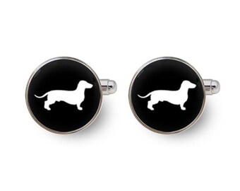 May include: A pair of round silver-toned cufflinks featuring a black circular face with a white silhouette of a dachshund dog in the center. The metal hardware has a polished finish and a standard toggle closure mechanism on the back.