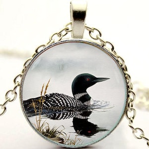 May include: A silver chain necklace with a round glass pendant featuring a black and white loon swimming in a lake. The loon has red eyes and is surrounded by water and reeds.
