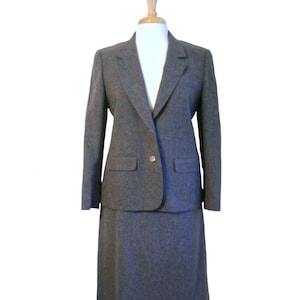 May include: A grey skirt suit on a mannequin. The suit comprises a blazer with a single button and two front pockets, and a matching knee-length skirt. The fabric looks like a textured wool blend. The mannequin is set against a white background.