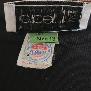 K&ouml;nnte beinhalten: Nahaufnahme von Kleidungsetiketten. Die Etiketten zeigen den Markennamen "eoele", Gr&ouml;&szlig;e 13 und "Made in U.S.A.". Die Etiketten sind an einem schwarzen Stoff befestigt.