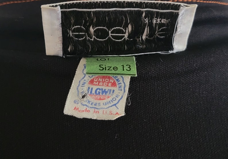 K&ouml;nnte beinhalten: Nahaufnahme von Kleidungsetiketten. Die Etiketten zeigen den Markennamen "eoele", Gr&ouml;&szlig;e 13 und "Made in U.S.A.". Die Etiketten sind an einem schwarzen Stoff befestigt.