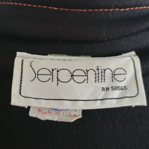 K&ouml;nnte beinhalten: Nahaufnahme eines schwarzen Kleidungsst&uuml;cks mit einem wei&szlig;en Etikett mit der Aufschrift "Serpentine" in einer stilisierten Schriftart. Das Etikett enth&auml;lt auch "RN 50565" und "Made in U.S.A.". Das Kleidungsst&uuml;ck hat orangefarbene N&auml;hte.