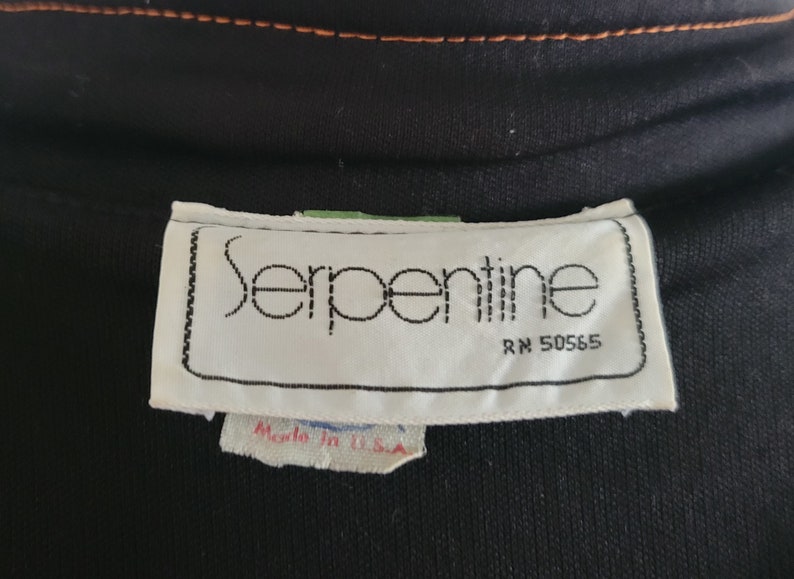K&ouml;nnte beinhalten: Nahaufnahme eines schwarzen Kleidungsst&uuml;cks mit einem wei&szlig;en Etikett mit der Aufschrift "Serpentine" in einer stilisierten Schriftart. Das Etikett enth&auml;lt auch "RN 50565" und "Made in U.S.A.". Das Kleidungsst&uuml;ck hat orangefarbene N&auml;hte.