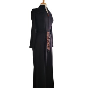 K&ouml;nnte beinhalten: Schwarzer Jumpsuit mit langen &Auml;rmeln und hohem Kragen, auf einer Schaufensterpuppe pr&auml;sentiert. Der Jumpsuit hat einen goldenen Rei&szlig;verschluss und einen gemusterten G&uuml;rtel. Das Design mit weitem Bein und das taillierte Oberteil schaffen eine elegante Silhouette.