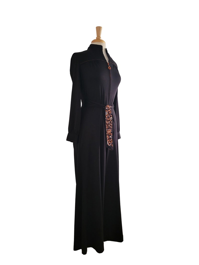 K&ouml;nnte beinhalten: Schwarzer Jumpsuit mit langen &Auml;rmeln und hohem Kragen, auf einer Schaufensterpuppe pr&auml;sentiert. Der Jumpsuit hat einen goldenen Rei&szlig;verschluss und einen gemusterten G&uuml;rtel. Das Design mit weitem Bein und das taillierte Oberteil schaffen eine elegante Silhouette.