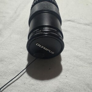 Puede incluir: Un objetivo de c&aacute;mara Olympus Quantaray de 49 mm negro con una tapa de objetivo. El objetivo tiene un agarre texturizado y est&aacute; sobre una superficie de color claro. La tapa del objetivo tiene el logotipo de Olympus.