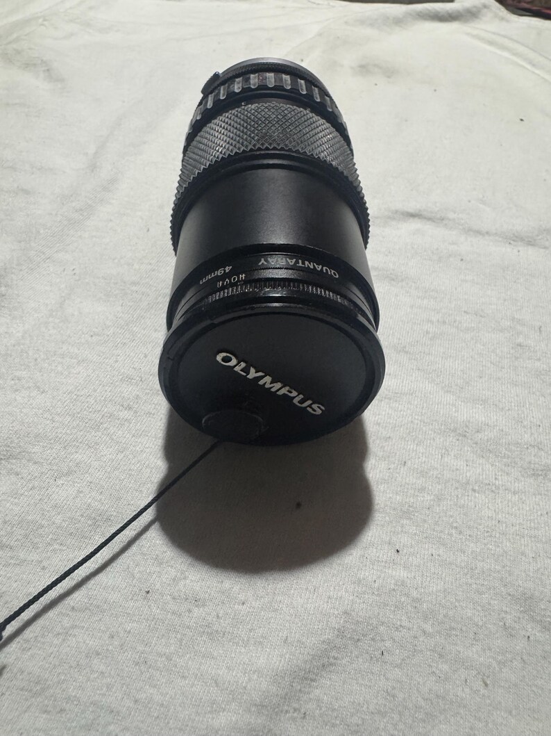 Puede incluir: Un objetivo de c&aacute;mara Olympus Quantaray de 49 mm negro con una tapa de objetivo. El objetivo tiene un agarre texturizado y est&aacute; sobre una superficie de color claro. La tapa del objetivo tiene el logotipo de Olympus.