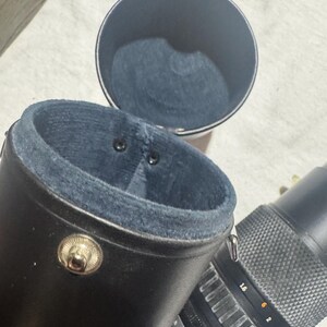 Puede incluir: Estuche negro de cuero para objetivo de c&aacute;mara con interior azul y cierre de bot&oacute;n plateado. El estuche est&aacute; abierto, revelando el forro azul. Un objetivo de c&aacute;mara es parcialmente visible junto al estuche.