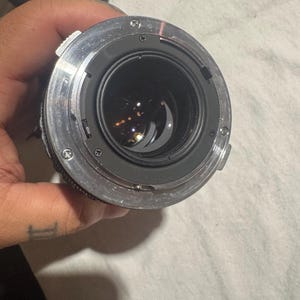 Puede incluir: Vista de cerca de una lente de c&aacute;mara, mostrando sus intrincados componentes internos. La lente presenta un exterior met&aacute;lico plateado con m&uacute;ltiples tornillos que aseguran su estructura. El cristal oscuro y reflectante de la lente es visible dentro del marco circular.
