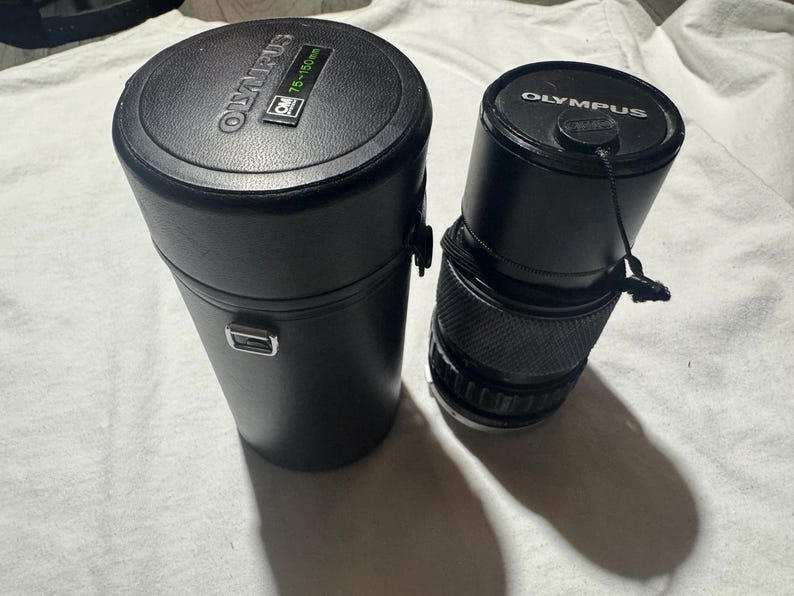 Puede incluir: Lente y estuche negros para c&aacute;mara Olympus. La lente es cil&iacute;ndrica con un agarre estriado y una tapa negra. El estuche es un cilindro alto y negro con un cierre plateado y el logotipo de Olympus. El estuche tiene una etiqueta verde que dice "75-150mm".