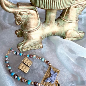 May include: An antique-style jewelry set featuring a beaded necklace and matching earrings with turquoise, brown, and gold beads. The set includes a pendant with a stylized figure and a small gold charm. A decorative bull-shaped stand is in the background.