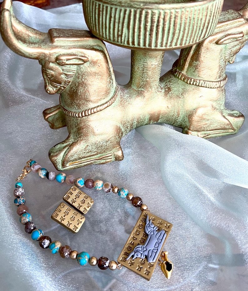 May include: An antique-style jewelry set featuring a beaded necklace and matching earrings with turquoise, brown, and gold beads. The set includes a pendant with a stylized figure and a small gold charm. A decorative bull-shaped stand is in the background.