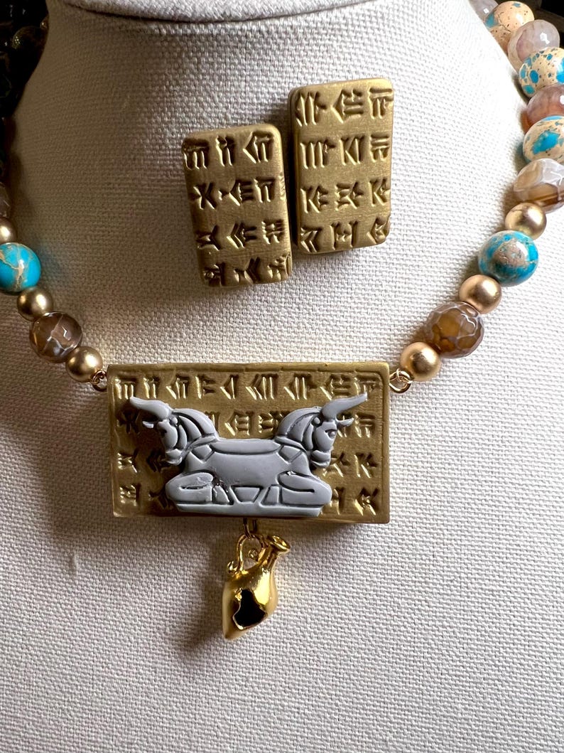 May include: A gold-toned necklace and matching earrings set. The necklace features a rectangular pendant with a gray winged creature and a small gold-toned charm. The earrings are rectangular with etched designs. The necklace has turquoise and brown beads.