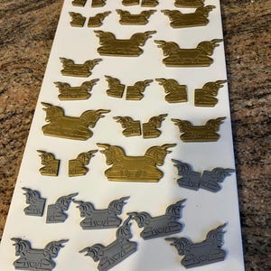 May include: A collection of gold and gray decorative horse-shaped embellishments arranged on a white surface. The embellishments vary in size and are likely for crafting or decorative purposes. The horses are in various poses.