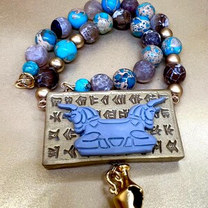 May include: A beaded necklace with a pendant featuring a blue bull design and gold accents. The necklace has a variety of colorful beads in shades of blue, brown, and gold. A small gold-colored vase hangs below the pendant.