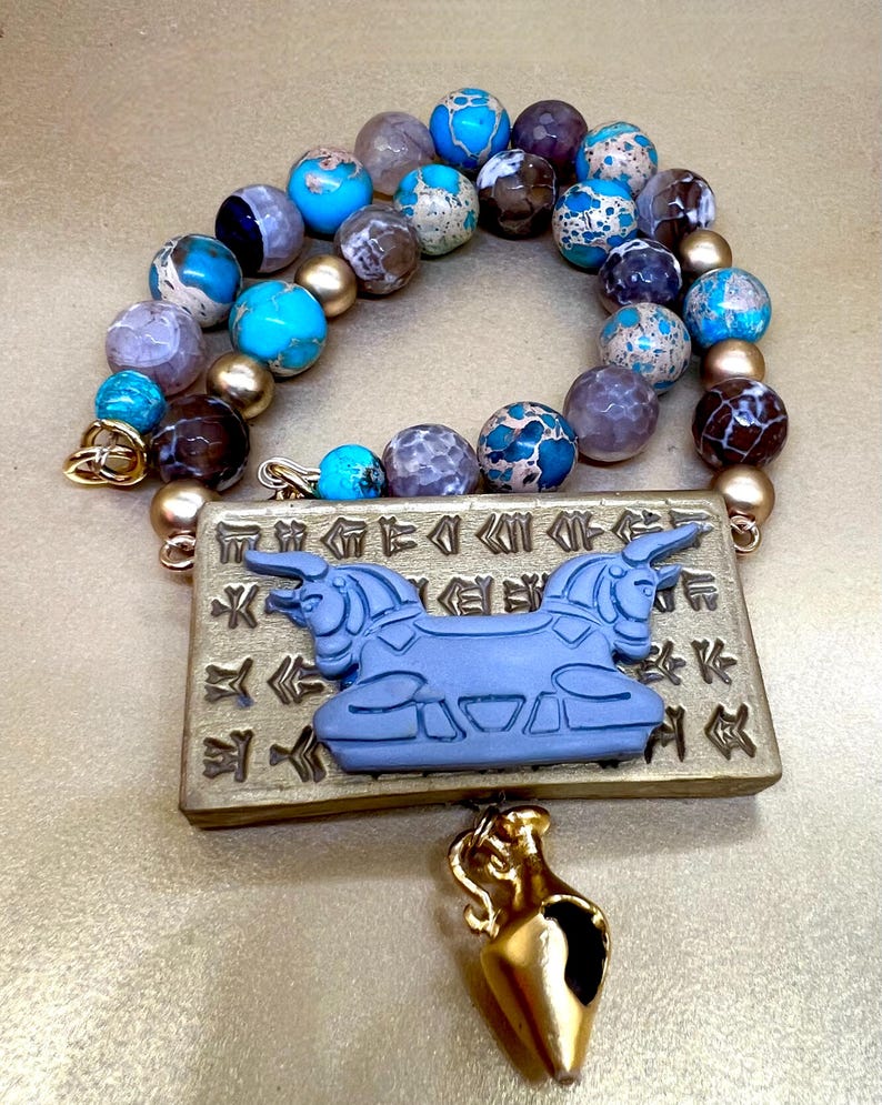 May include: A beaded necklace with a pendant featuring a blue bull design and gold accents. The necklace has a variety of colorful beads in shades of blue, brown, and gold. A small gold-colored vase hangs below the pendant.