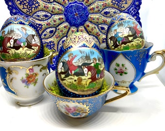 May include: Ornate, hand-painted eggs with miniature scenes of figures on horseback, displayed in decorative teacups. The eggs and cups feature blue, gold, and floral designs. A matching blue plate is in the background.
