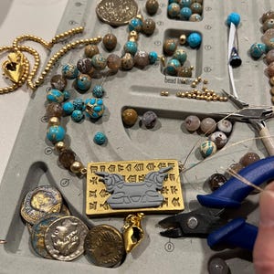 May include: A jewelry-making workspace with various beads, gold-colored chains, and tools. The beads are in shades of blue, brown, and gold. A necklace with a rectangular pendant featuring a gray bull design is visible, along with several antique coins.