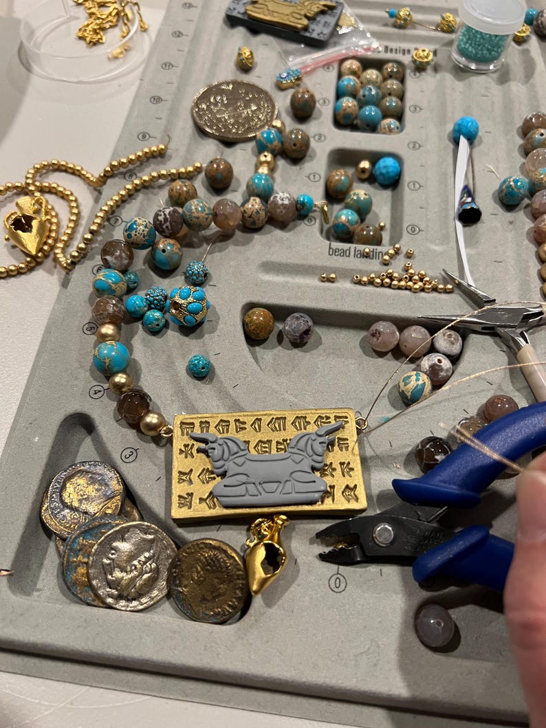 May include: A jewelry-making workspace with various beads, gold-colored chains, and tools. The beads are in shades of blue, brown, and gold. A necklace with a rectangular pendant featuring a gray bull design is visible, along with several antique coins.