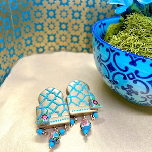 May include: A pair of ornate earrings with a light grey base, accented with turquoise blue lines and floral details. The earrings have a dangling bead design with turquoise and floral accents. The background features a blue and gold patterned surface.