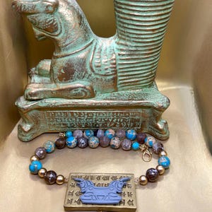 May include: A beaded bracelet with blue, brown, and gold beads, and a rectangular pendant with a blue bull design. A gold-colored charm hangs below the pendant. The bracelet is displayed in front of a bronze-colored sculpture.