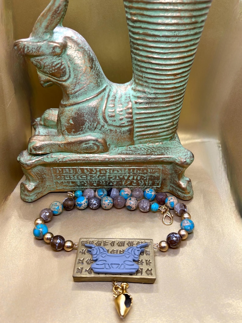 May include: A beaded bracelet with blue, brown, and gold beads, and a rectangular pendant with a blue bull design. A gold-colored charm hangs below the pendant. The bracelet is displayed in front of a bronze-colored sculpture.