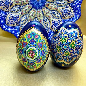 May include: Two navy blue, hand-painted decorative eggs with intricate floral and geometric designs in gold, yellow, pink, and green. A matching blue plate with similar patterns is in the background. The eggs are a unique, artistic home decor item.