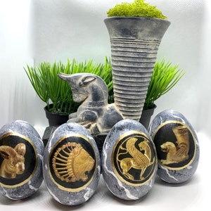 Può includere: Uova decorative grigie e dorate con disegni a rilievo di animali. Ogni uovo presenta un animale diverso, tra cui un ariete, un leone, un grifone e una creatura simile a un leone. Le uova sono presentate con verde e un vaso decorativo.