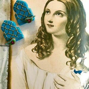 Può includere: Due oggetti decorativi turchese e dorati a forma di pianoforte con accenti floreali sono esposti su una stampa vintage di una donna. La stampa è incorniciata in una cornice azzurra e grigia invecchiata. La donna nella stampa tiene in mano un uccello.