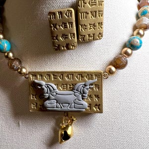May include: A gold-toned necklace and matching earrings set. The necklace features a rectangular pendant with a gray winged creature and a small gold-toned charm. The earrings are rectangular with etched designs. The necklace has turquoise and brown beads.