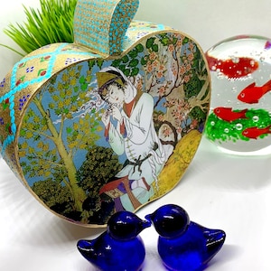 May include: An ornate, heart-shaped box with a painted scene of a person in traditional clothing, surrounded by trees and flowers. The box has gold and turquoise accents. Two small, blue glass bird figurines and a glass paperweight with red fish are also present.