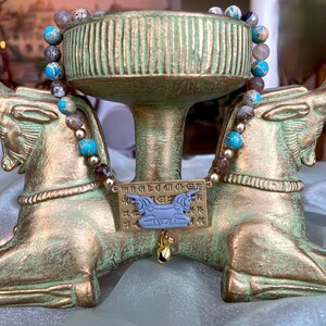 May include: A decorative gold-toned stand with bull-shaped supports, holding a beaded necklace with blue and brown stones. A rectangular plaque with hieroglyphs and a blue bull pendant hangs below the necklace. The stand has a weathered, antique appearance.