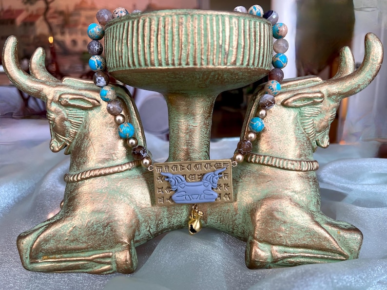 May include: A decorative gold-toned stand with bull-shaped supports, holding a beaded necklace with blue and brown stones. A rectangular plaque with hieroglyphs and a blue bull pendant hangs below the necklace. The stand has a weathered, antique appearance.