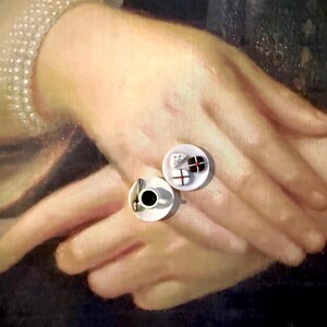 May include: Miniature coffee cup with a spoon and a small plate with a few chocolates and cheese. The items are set against a backdrop of a painting featuring hands and a pearl necklace.