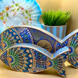 May include: Two decorative wooden fish sculptures with intricate blue, gold, and green mosaic patterns. The fish are in front of a blue and white ruffled bowl and a small potted plant with green grass.