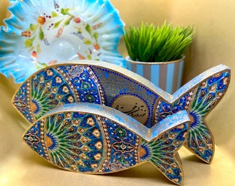 May include: Two decorative wooden fish sculptures with intricate blue, gold, and green mosaic patterns. The fish are in front of a blue and white ruffled bowl and a small potted plant with green grass.