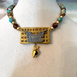 May include: A beaded necklace with a gold pendant. The necklace features turquoise, brown, and gold beads. The gold pendant has a rectangular shape with an engraved design and a small gold charm hanging below.