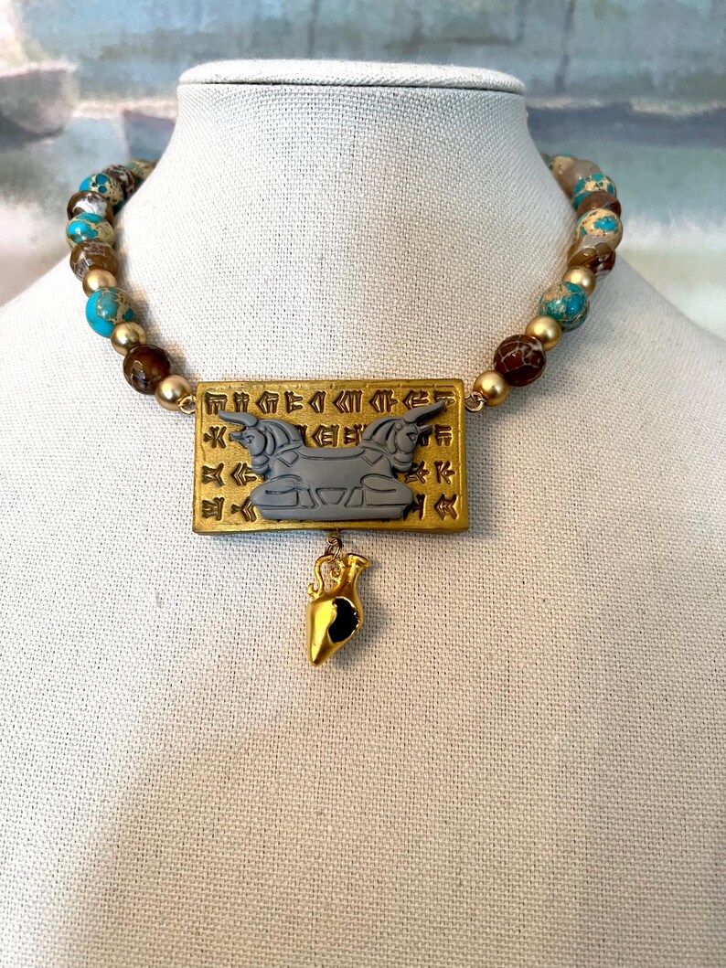 May include: A beaded necklace with a gold pendant. The necklace features turquoise, brown, and gold beads. The gold pendant has a rectangular shape with an engraved design and a small gold charm hanging below.