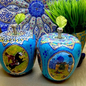 Op de afbeelding: Twee turquoise appelvormige ornamenten met gedetailleerde patronen en gouden accenten. Ze tonen miniatuurschilderijen van ruiters en architectuurscènes. Elke appel heeft een glinsterende steel en een groen blad.