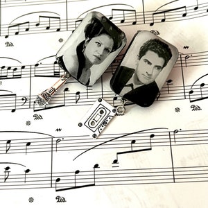 May include: Two rectangular pendants with black and white portraits of people, one with a microphone charm and the other with a cassette tape charm, rest on sheet music. The background is a white sheet of music with black notes and the text "J. = 57" and "Red."