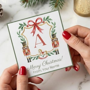 Può includere: Un biglietto di Natale con un design ad acquerello. Il biglietto presenta un fiocco rosso, una ghirlanda verde e la lettera "A". Il biglietto ha anche due figure di schiaccianoci e il testo "Merry Christmas! From, Your Name".