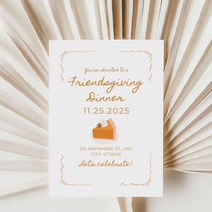 Puede incluir: Una tarjeta de invitación blanca con el texto "You're invited to a Friendgiving Dinner 11.25.2025" y una ilustración de tarta de calabaza. La tarjeta está colocada sobre un fondo de hojas de palma secas.