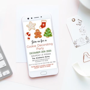 May include: A smartphone displays a Christmas cookie decorating party invitation. The invitation features gingerbread men, Christmas trees, and stockings. The phone is on a white surface with a keyboard, flowers, and a cup of coffee.