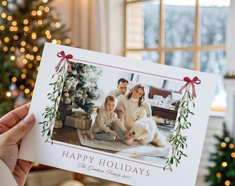 May include: A person holds a Christmas card with a family photo and the text "HAPPY HOLIDAYS." The card has a red border with green and red accents. A decorated Christmas tree and gifts are in the background.