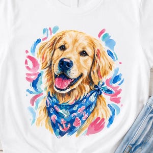 May include: White t-shirt featuring a watercolor illustration of a Golden Retriever wearing a blue bandana with anchor and seashell designs. The dog is smiling, surrounded by pink and blue brushstrokes. The shirt is paired with blue jeans.