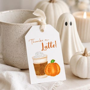 May include: A white tag with the text "Thanks a Latte!" features a watercolor illustration of a latte and a pumpkin. The tag is tied to a beige mug with a cream-colored ribbon. White pumpkins, a ghost figurine, and a candle complete the autumn-themed display.