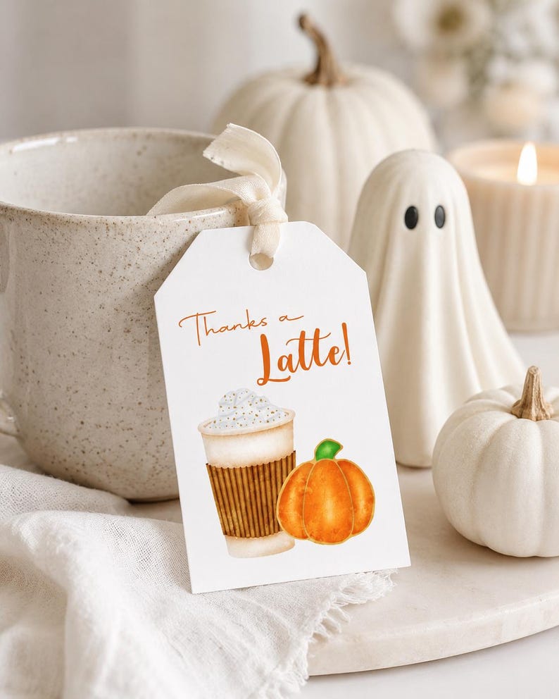May include: A white tag with the text "Thanks a Latte!" features a watercolor illustration of a latte and a pumpkin. The tag is tied to a beige mug with a cream-colored ribbon. White pumpkins, a ghost figurine, and a candle complete the autumn-themed display.